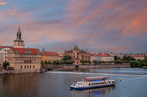 Photo of Prague at dusk with the Old Town Water Tower in the background and a boat gently sailing along the Vlatava River.