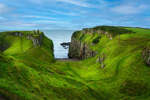 Rolling green cliffs in Ireland with Dunseverick Castle in the backgrounds with sheep in the hills. There is a view of the rugged coastline with the Atlantic Ocean in the background.