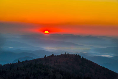 Sunrise in the mountqains of North Carolina off of the Blue Ridge Parkway. The sky is golden wirth the horizon deep orange. There are layers of blue clouds below a mountain top.