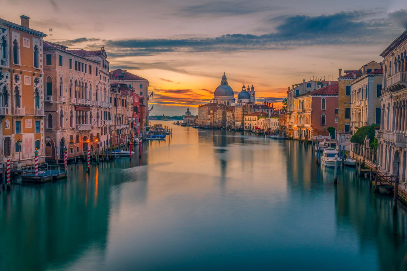 The Grand Canal in Venice at sunrise with a stillness in the water and hues of orange and yellow in the sky.