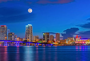 A cityscape of downtown Miami and the bridge connecting Watson Island to downtown. This was taken during the blue hour and the sky is a deep blue color.  There is a full moon over Miami and the city lights are on and colorful.