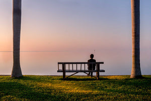 A woman sittling on a bench at sunrise at the edge of the water at Biscayne Bay. The woman is sitting on a bench that is between two palm trees and the sky is  pale pinks and purples. 